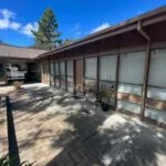 Single-story medical clinic building with brown wooden exterior, large windows with white blinds, and brick patio walkway on a sunny day