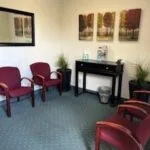 Waiting room with burgundy upholstered chairs, black console table, framed artwork on cream walls, and potted plants on blue patterned carpet