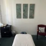 Massage or treatment room with white draped table, black nightstand, burgundy cabinet, and three framed botanical prints on white wall
