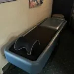Massage table with black top and blue base in clinic room with beige walls and posters