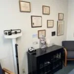 Office interior with black cabinet, framed certificates on wall, and medical equipment on white shelf