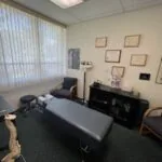 Chiropractic treatment room with massage table, black cabinet, wall-mounted light, framed certificates on wall, and windows with blinds.