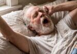 Older man with gray beard stretching in bed with arms raised after waking up